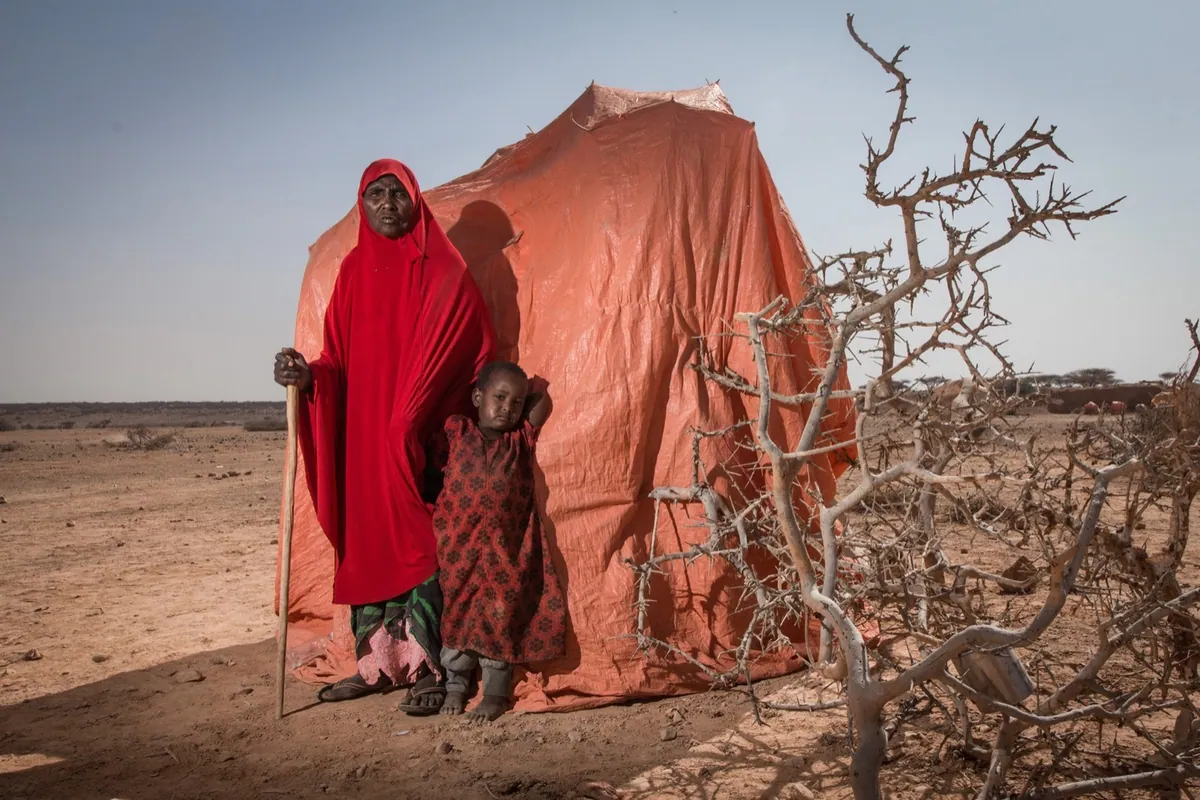 BBC, photo 06. Grandmother Amina stands with her daughter in front of a makeshift shelter in an informal settlement outside Yogoori.
